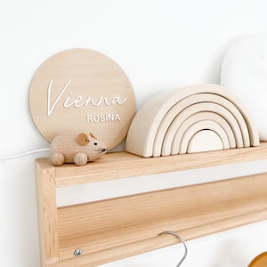 May include: A wooden shelf with a wooden rainbow toy, a wooden mouse toy, and a round wooden sign with the name "Vienna Rosina" written in white.