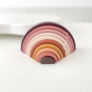 May include: A wooden rainbow stacking toy with a variety of pink, orange, and brown shades. The toy is made of 7 pieces that can be stacked on top of each other.