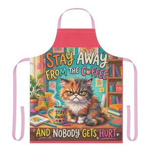 May include: A pink-trimmed apron featuring a graphic of a grumpy calico cat sitting on a desk next to a steaming mug. The text reads STAY AWAY FROM THE COFFEE AND NOBODY GETS HURT. The background shows a bookshelf, a potted plant, and art supplies.
