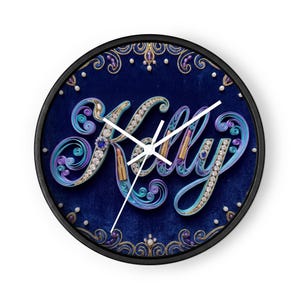 Può includere: Un orologio da parete rotondo con cornice nera e sfondo in velluto blu intenso. Il nome Kelly è scritto in un carattere stilizzato realizzato con carta quilling azzurra e viola, arricchito da strass incastonati e piccoli rossetti.