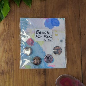 Puede incluir: Un paquete de cuatro pines de escarabajos sobre un fondo colorido de acuarela. Los pines son redondos con varios diseños de escarabajos. El embalaje incluye el texto "Beetle Pin Pack 4x Pins". Los pines se muestran sobre una superficie de madera.