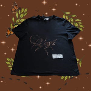 Peut inclure: Un t-shirt noir à manches courtes avec un motif d'araignée marron imprimé au centre de la poitrine. Un petit écusson rectangulaire en tissu clair aux bords effilochés est cousu sur le côté inférieur droit du devant du t-shirt.