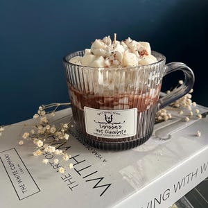 Könnte beinhalten: Eine Kerze in Form einer Glas-Kaffeetasse mit einem braunen und weißen Heißschokoladen-Duft. Die Kerze ist mit weißer Schlagsahne und braunen Streuseln verziert. Die Kerze steht auf einem weißen Buch mit dem Text "The Art of Living with" auf dem Einband.