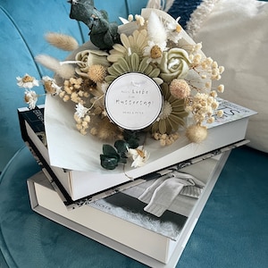 Puede incluir: Un ramo de flores secas en tonos blancos y verdes, atado con una cinta blanca. Una etiqueta redonda con el texto "Alles Liebe zum Muttertag! Heike & Peter" está unida al ramo.