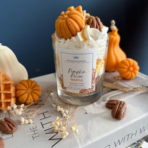 Könnte beinhalten: Ein Duftkerze im klaren Glas mit Kürbis-Pekan-Waffel-Duft, dekoriert mit geschlagener Wachs, orangefarbenen Kürbissen und Pekannüssen. Die Kerze steht auf einem Buch mit der Aufschrift "Living With" und "The Art Of".