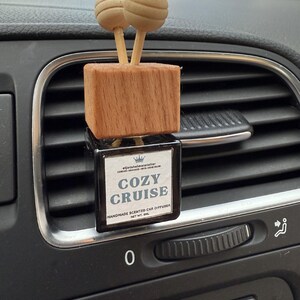 Könnte beinhalten: Ein Auto-Lufterfrischer mit einem Holzblock und zwei kugelförmigen Holzaufsätzen. Auf der schwarzen rechteckigen Flasche steht "COZY CRUISE". Der Lufterfrischer ist an einer Lüftungsöffnung im Auto befestigt.