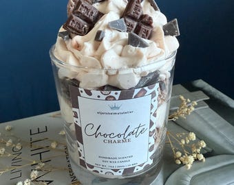Puede incluir: Una vela de vidrio con aroma a chocolate y vainilla. La vela está cubierta con crema batida y trozos de chocolate, incluyendo una barra de chocolate y una galleta de chocolate. La etiqueta dice "Chocolate Charme" y "Vela de cera de soja perfumada hecha a mano, Peso neto 7 oz (200g) 50 horas."