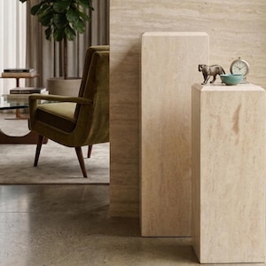 May include: Two rectangular beige travertine stone pedestals with a natural veined texture. The shorter pedestal displays a small bronze-toned panther figurine, a round teal ceramic bowl, and a silver-toned analog clock with a white face.