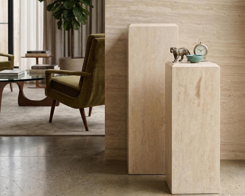 May include: Two rectangular beige travertine stone pedestals with a natural veined texture. The shorter pedestal displays a small bronze-toned panther figurine, a round teal ceramic bowl, and a silver-toned analog clock with a white face.