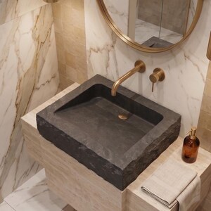 Pu&ograve; includere: Un moderno lavabo da bagno in pietra grigio scuro. Il bacino rettangolare poggia su un piano in pietra di colore chiaro. Un rubinetto dorato e accessori abbinati sono montati sulla parete sopra il lavabo. Sono visibili anche uno specchio rotondo e un lampadario.