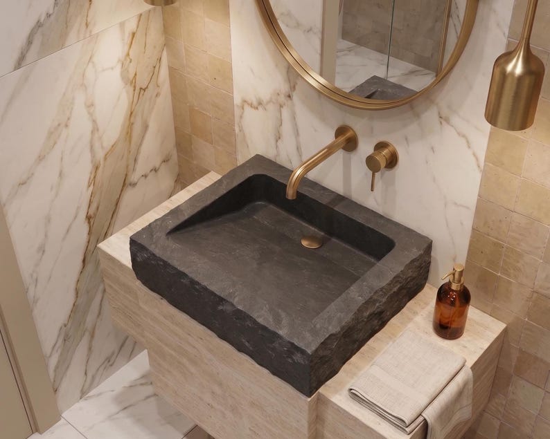 Pu&ograve; includere: Un moderno lavabo da bagno in pietra grigio scuro. Il bacino rettangolare poggia su un piano in pietra di colore chiaro. Un rubinetto dorato e accessori abbinati sono montati sulla parete sopra il lavabo. Sono visibili anche uno specchio rotondo e un lampadario.