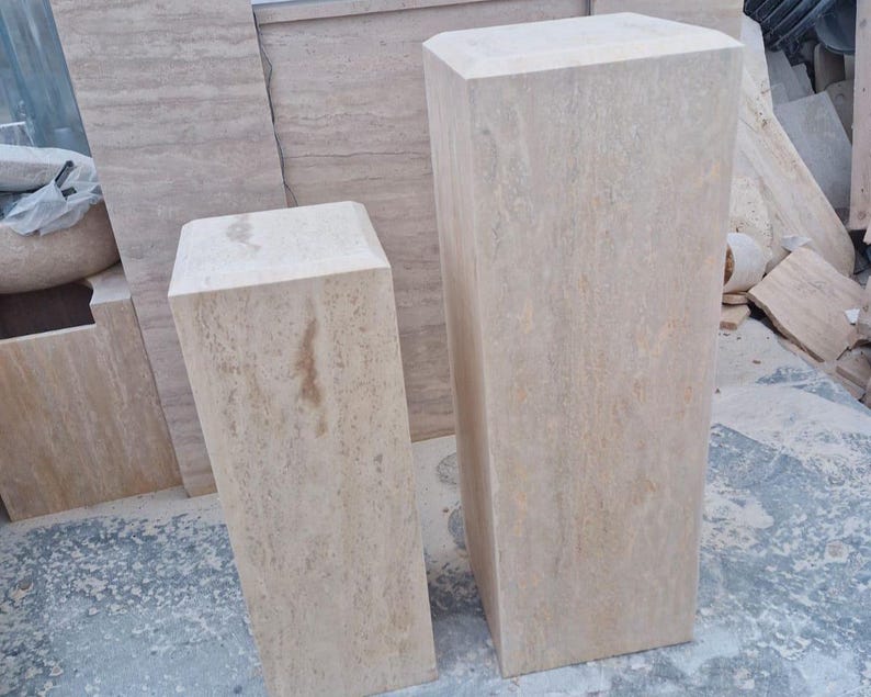 May include: Two rectangular pedestals crafted from light beige travertine stone with a natural porous texture and visible veining. The pedestals feature flat tops with beveled edges and vertical sides, standing upright as solid, block-like display columns.