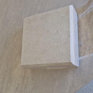 May include: A square, light beige stone tile with a smooth, matte surface and a beveled edge. The tile features a natural, mottled texture with subtle variations in color throughout the material, showcasing a clean, geometric shape and finish.