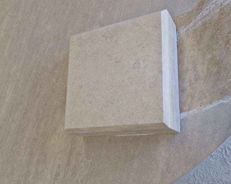 May include: A square, light beige stone tile with a smooth, matte surface and a beveled edge. The tile features a natural, mottled texture with subtle variations in color throughout the material, showcasing a clean, geometric shape and finish.
