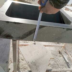 Pu&ograve; includere: Un lavandino rettangolare in pietra grigio scuro con angoli arrotondati. Un metro a nastro &egrave; tenuto sul lato del lavandino. Il lavandino &egrave; posto su un supporto metallico. Il lavandino &egrave; in pietra ruvida e strutturata.