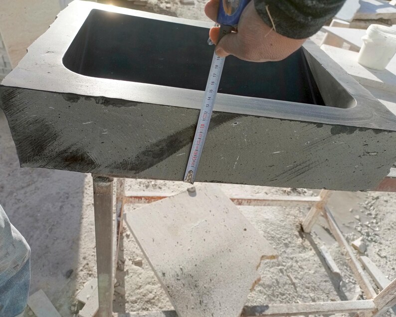 Pu&ograve; includere: Un lavandino rettangolare in pietra grigio scuro con angoli arrotondati. Un metro a nastro &egrave; tenuto sul lato del lavandino. Il lavandino &egrave; posto su un supporto metallico. Il lavandino &egrave; in pietra ruvida e strutturata.