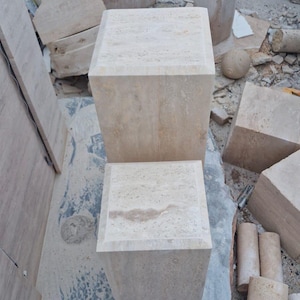 May include: Two square, beige travertine stone pedestals with a natural porous texture and beveled top edges. The stone blocks feature light tan and cream color variations throughout their surfaces, showcasing the organic patterns of the natural material.