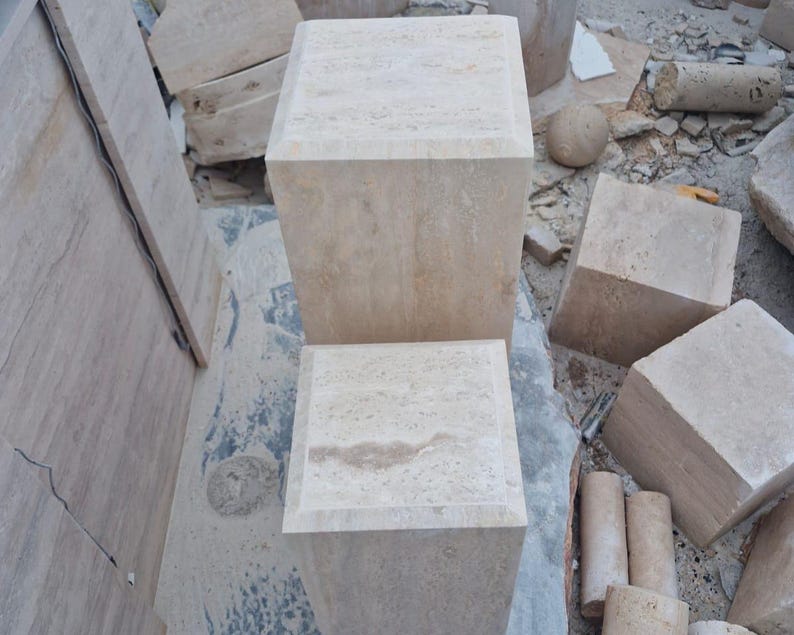 May include: Two square, beige travertine stone pedestals with a natural porous texture and beveled top edges. The stone blocks feature light tan and cream color variations throughout their surfaces, showcasing the organic patterns of the natural material.