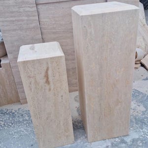 May include: Two rectangular pedestals made of beige travertine stone with natural veining and smooth surfaces. The taller pedestal stands to the right, while the shorter pedestal is positioned to the left, both featuring beveled top edges.