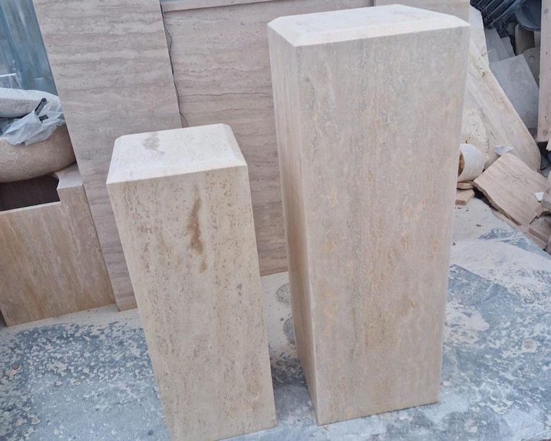 May include: Two rectangular pedestals made of beige travertine stone with natural veining and smooth surfaces. The taller pedestal stands to the right, while the shorter pedestal is positioned to the left, both featuring beveled top edges.