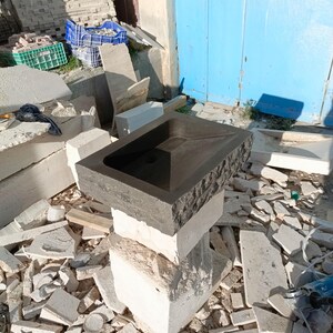 Pu&ograve; includere: Un lavabo rettangolare in pietra grigio scuro con esterno strutturato, appoggiato su una pila di blocchi di pietra colorati. Il lavabo ha uno scarico centrale e un interno liscio. Lo sfondo mostra materiali da costruzione e una porta blu.