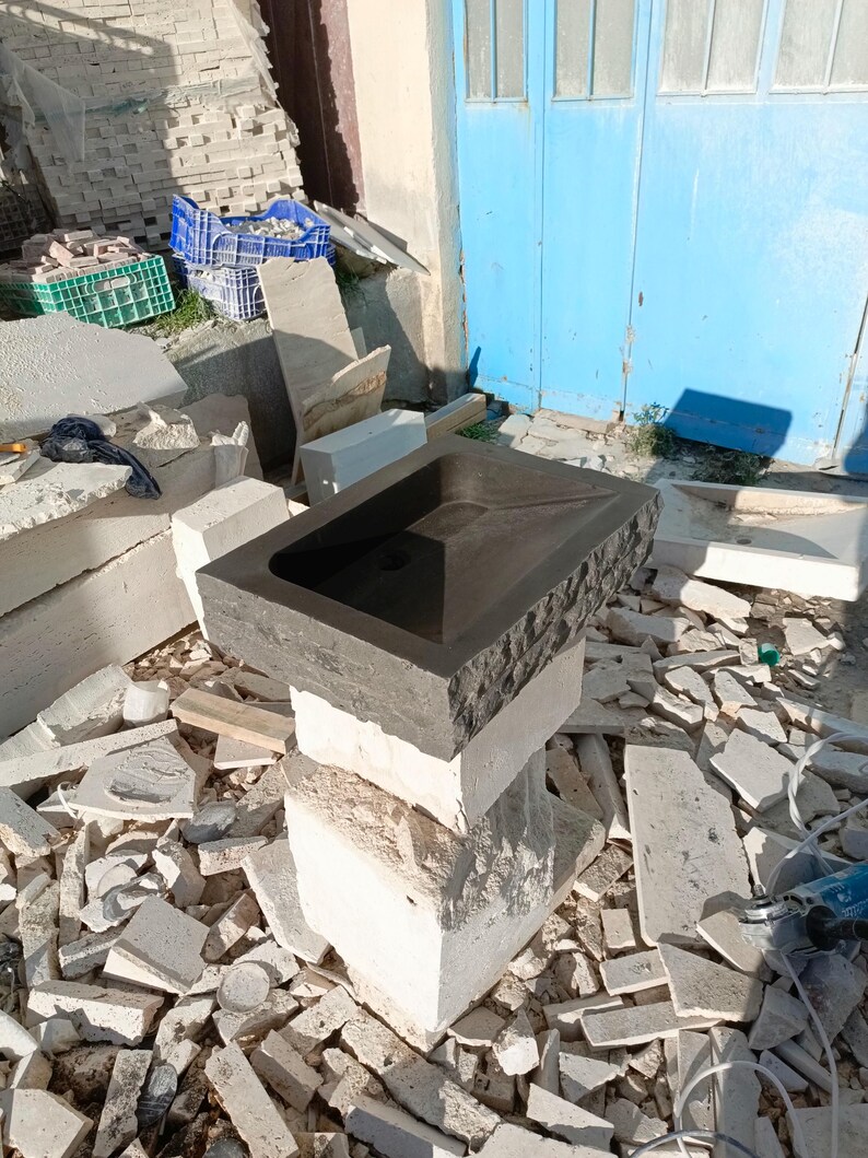 Pu&ograve; includere: Un lavabo rettangolare in pietra grigio scuro con esterno strutturato, appoggiato su una pila di blocchi di pietra colorati. Il lavabo ha uno scarico centrale e un interno liscio. Lo sfondo mostra materiali da costruzione e una porta blu.