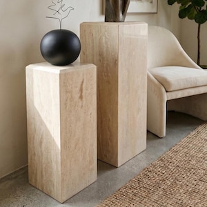 May include: Two rectangular pedestals crafted from beige travertine stone with a natural porous texture. The shorter pedestal on the left supports a smooth, matte black spherical object topped with a thin, dark wire sculpture of a bird.