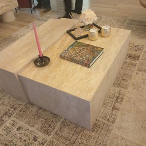 Könnte beinhalten: Ein hellbeige rechteckiger Couchtisch mit einem Buch, einer Kerze und dekorativen Gegenständen. Der Tisch hat ein geteiltes Design und steht auf einem gemusterten Teppich. Eine rosa Kerze befindet sich in einem schwarzen Halter.