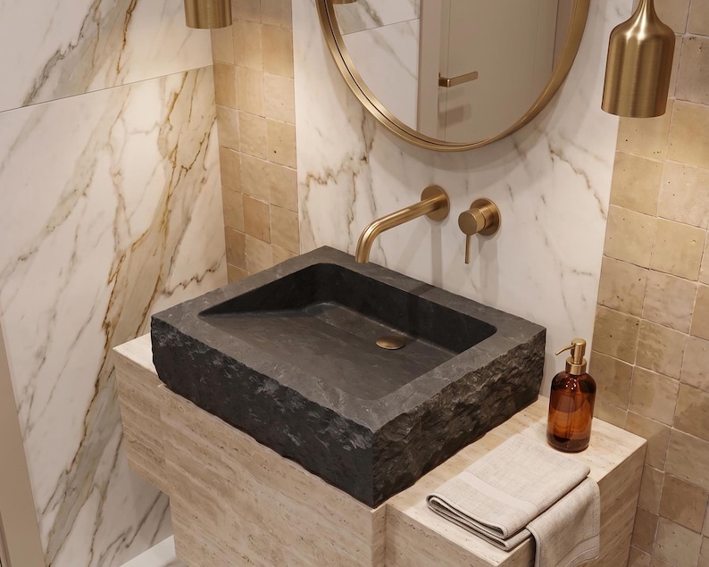 Pu&ograve; includere: Un lavabo rettangolare in pietra grigio scuro con un rubinetto in ottone e un dosatore di sapone abbinato. Il lavabo &egrave; appoggiato su un piano di lavoro in pietra colorata con un asciugamano beige piegato. Il bagno ha pareti in marmo e piastrelle.