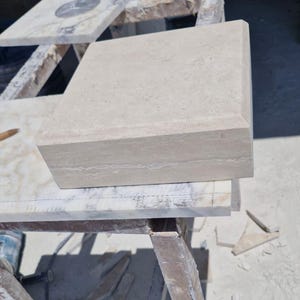 May include: A square, beige stone block with a smooth, flat top surface and beveled edges. The stone features a natural, porous texture with subtle horizontal veining visible along the side profile, showcasing a solid, architectural construction.