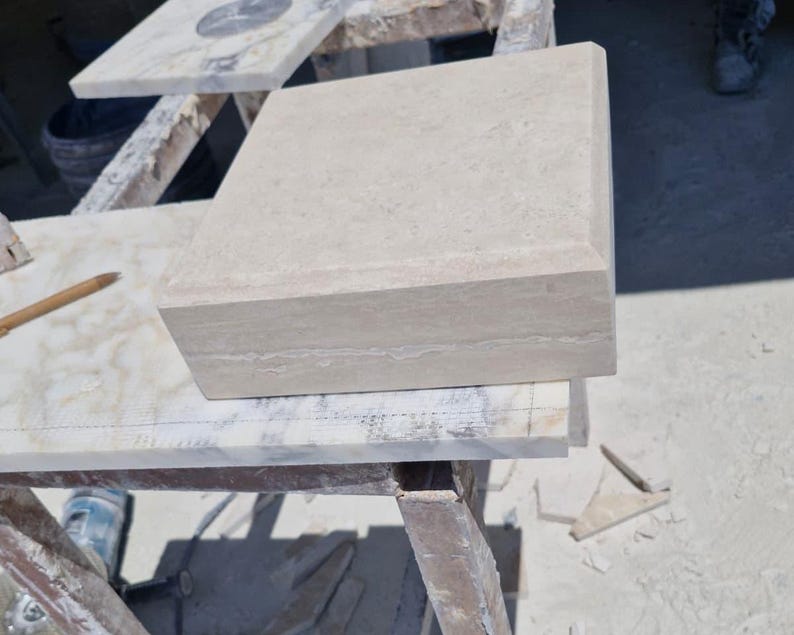 May include: A square, beige stone block with a smooth, flat top surface and beveled edges. The stone features a natural, porous texture with subtle horizontal veining visible along the side profile, showcasing a solid, architectural construction.