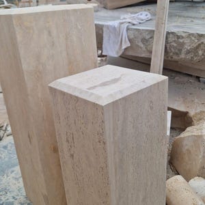 May include: Two rectangular beige travertine stone pedestals with a porous, natural texture and beveled top edges. The taller pedestal stands behind the shorter one, both displaying the characteristic light tan color and organic veining of stone.