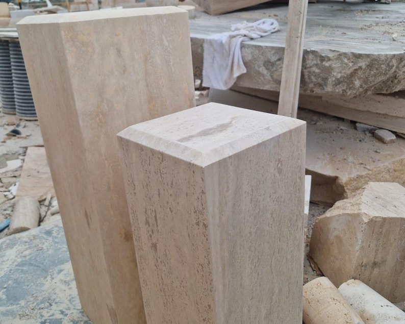 May include: Two rectangular beige travertine stone pedestals with a porous, natural texture and beveled top edges. The taller pedestal stands behind the shorter one, both displaying the characteristic light tan color and organic veining of stone.