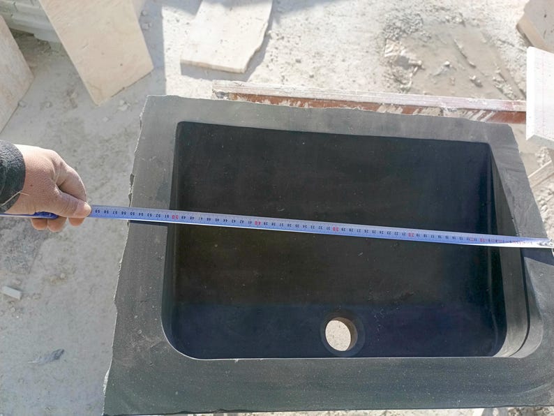 Pu&ograve; includere: Un lavandino rettangolare in pietra grigio scuro con interno arrotondato. Un metro a nastro &egrave; posto sopra il lavandino, indicando le sue dimensioni. Il lavandino ha un foro di scarico ed &egrave; inserito in una cornice di pietra grezza.