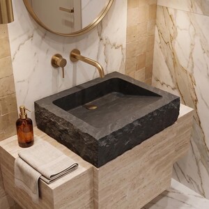Pu&ograve; includere: Un lavabo moderno per il bagno in pietra grigio scuro. Il lavabo rettangolare &egrave; appoggiato su un mobile di colore chiaro. Un rubinetto e uno specchio dorati sono montati a parete. Un dosatore di sapone marrone e un asciugamano piegato sono sul mobile.