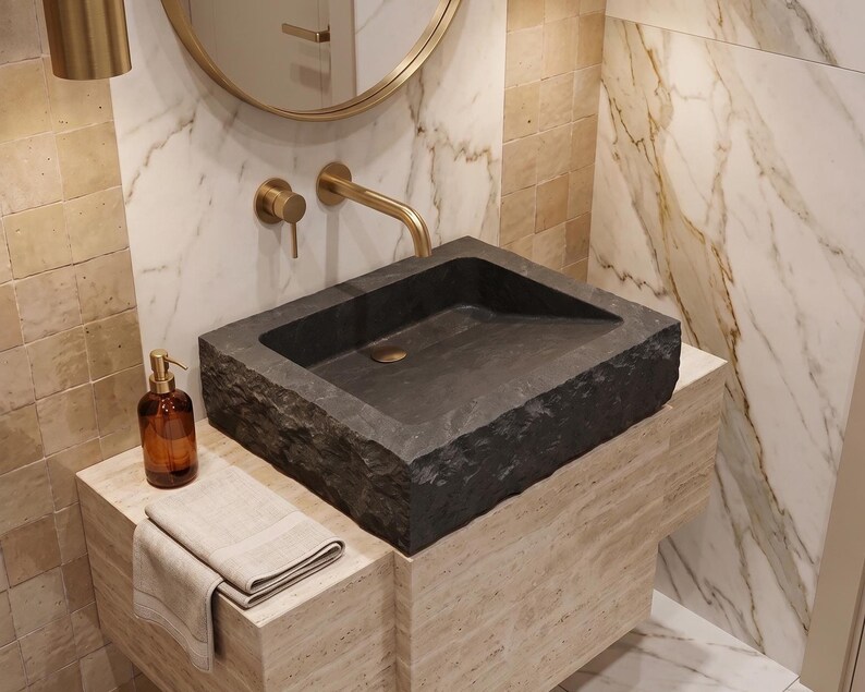 Pu&ograve; includere: Un lavabo moderno per il bagno in pietra grigio scuro. Il lavabo rettangolare &egrave; appoggiato su un mobile di colore chiaro. Un rubinetto e uno specchio dorati sono montati a parete. Un dosatore di sapone marrone e un asciugamano piegato sono sul mobile.