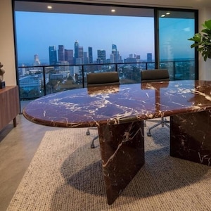 May include: An oval conference table made of deep red marble with prominent white veining, supported by two thick, matching marble slab pedestals. The table surface is polished and reflective, positioned in a room with a view of a city skyline.