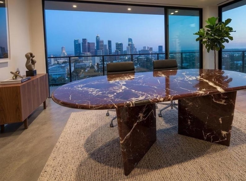 May include: An oval conference table made of deep red marble with prominent white veining, supported by two thick, matching marble slab pedestals. The table surface is polished and reflective, positioned in a room with a view of a city skyline.