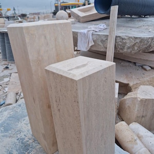 May include: Two rectangular stone pedestals with a light beige, porous texture and beveled top edges. The taller pedestal stands to the left, while the shorter pedestal is positioned in the foreground to the right, both featuring a natural finish.