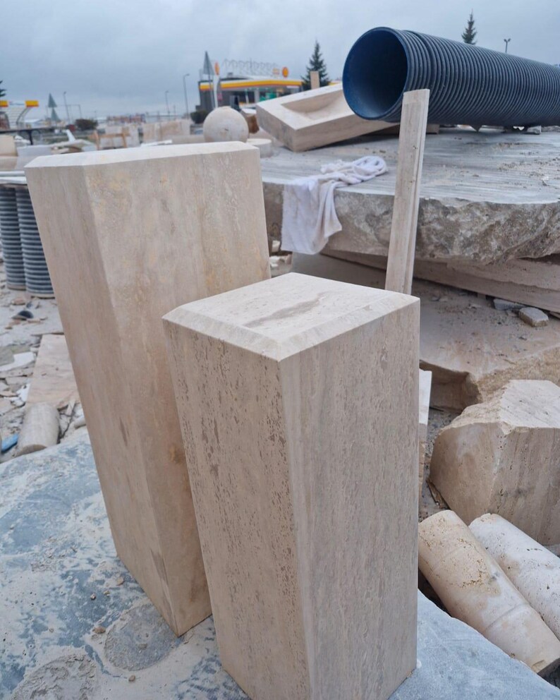May include: Two rectangular stone pedestals with a light beige, porous texture and beveled top edges. The taller pedestal stands to the left, while the shorter pedestal is positioned in the foreground to the right, both featuring a natural finish.