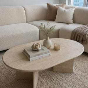 Pode incluir: Uma mesa de centro oval de travertino bege com base de pedestal. Sobre a mesa estão dois livros empilhados intitulados Live Beautifully e Kinfolk, uma escultura de nó bege, um pequeno vaso de cerâmica bege com ramos e uma vela de vidro.