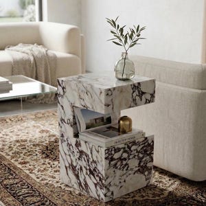 Könnte beinhalten: Ein Beistelltisch aus Marmor mit einem einzigartigen C-förmigen Design. Der Tisch hat ein weißes und burgunderfarbenes Adernmuster. Eine Glasvase mit Grün und ein kleiner goldfarbener Gegenstand stehen oben. Bücher werden im offenen Raum des Tisches ausgestellt.
