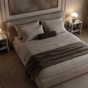 Può includere: Una struttura letto imbottita color beige con testiera abbinata, vestita con biancheria da letto beige chiaro, due cuscini rettangolari e un cuscino cilindrico marrone scuro. Una coperta marrone scuro è drappeggiata ai piedi del letto.