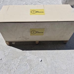 May include: A rectangular light-colored wooden crate with a flat top and a base featuring small support blocks. The crate displays two yellow hexagonal labels, one on the top and one on the front, each featuring the text NIZA DEKORASYON.