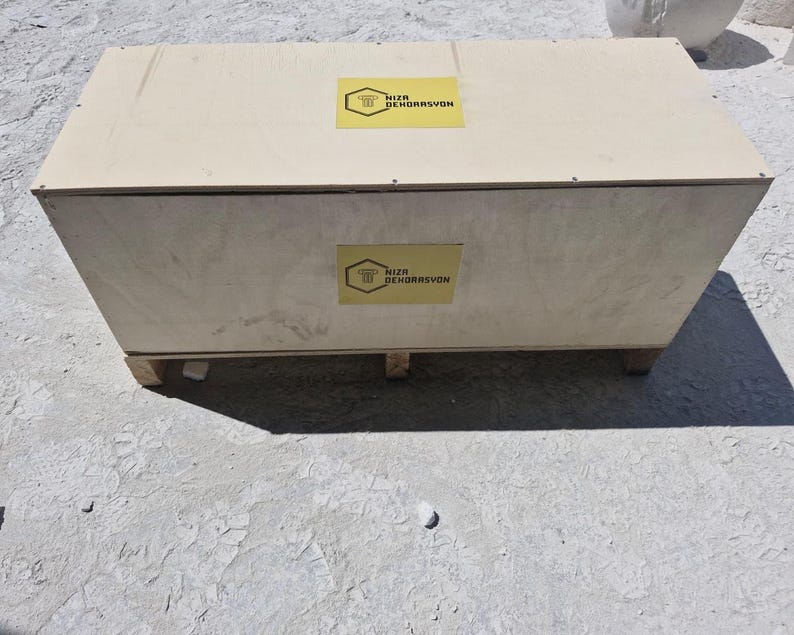 May include: A rectangular light-colored wooden crate with a flat top and a base featuring small support blocks. The crate displays two yellow hexagonal labels, one on the top and one on the front, each featuring the text NIZA DEKORASYON.