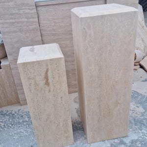 May include: Two rectangular pedestals crafted from beige travertine stone with natural veining and porous textures. The pedestals feature flat tops and vertical sides, standing upright as solid blocks with a light tan and cream color palette.