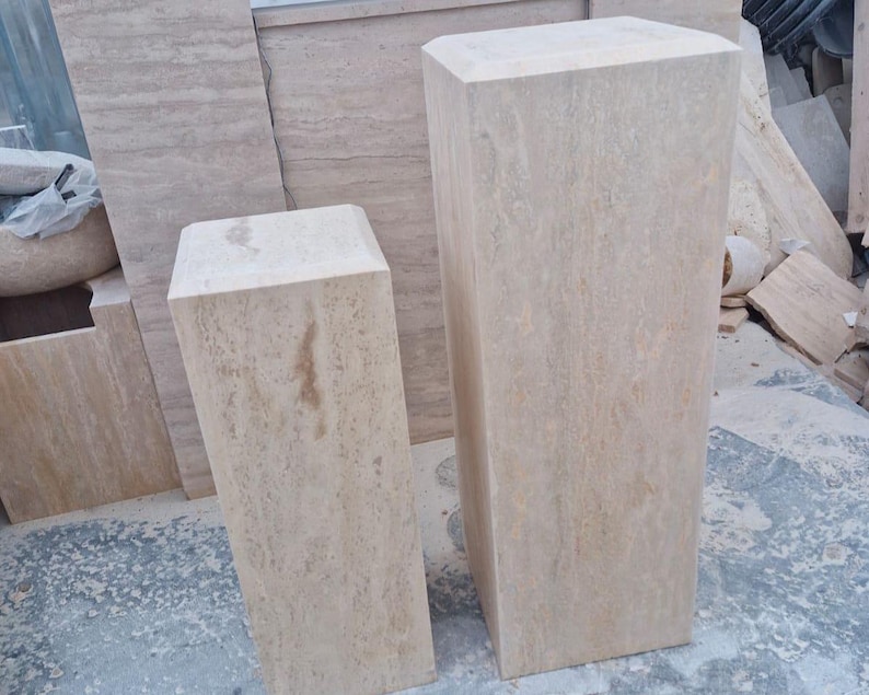 May include: Two rectangular pedestals crafted from beige travertine stone with natural veining and porous textures. The pedestals feature flat tops and vertical sides, standing upright as solid blocks with a light tan and cream color palette.