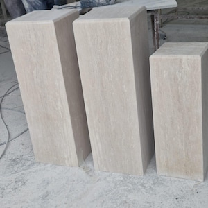 May include: Three rectangular stone pedestals in a light beige color with natural veining patterns. Each pedestal features a smooth, flat top surface and vertical sides, standing upright in a workshop setting with a consistent stone texture.