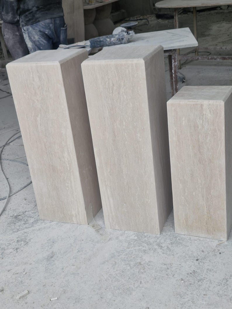 May include: Three rectangular stone pedestals in a light beige color with natural veining patterns. Each pedestal features a smooth, flat top surface and vertical sides, standing upright in a workshop setting with a consistent stone texture.