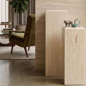 May include: Two rectangular beige travertine stone pedestals of varying heights. The shorter pedestal holds a small bronze-colored tiger figurine, a small teal bowl, and a silver-toned analog clock with a white face and black hands.